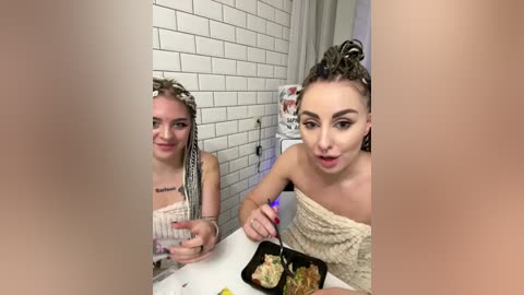 Video of two young women, one with dreadlocks and the other with braids, seated at a white table in a bathroom. They are casually dressed and smiling.