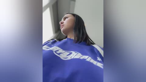 Media: A video of a young woman with straight, shoulder-length black hair, wearing a blue and white sweatshirt with \"Spartans\" in bold white letters, gazing upward with a contemplative expression. The background is blurred, featuring a white wall and a window.
