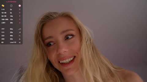 A video of a smiling, fair-skinned woman with long, wavy blonde hair, wearing a grey sweater, against a plain, light grey background. The image includes a digital display showing a calendar and weather information.