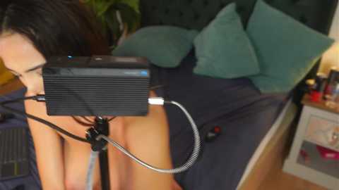 Video of a woman with dark hair, nude, sitting on a bed with teal pillows and a green blanket, using a black speaker on a stand.