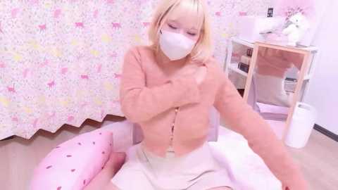 A video shows a blonde woman with a white mask, wearing a light pink cardigan, seated on a pink polka-dotted pillow, sewing in a room with floral wallpaper and a sewing machine.