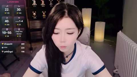 Media: Video of an East Asian woman with long black hair, wearing a white t-shirt, sitting in a dimly lit room with a TV displaying game stats, a radiator, and a plant.