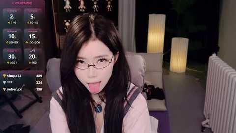 Media: Video of a young Asian woman with long black hair, glasses, and a playful expression, sitting on a couch in a dimly lit living room with two tall, lit lamps and a TV displaying game stats.