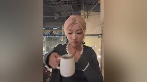 Video of an Asian woman with fair skin, light pink headscarf, and black dress with white trim, holding a white mug. Background shows a dimly lit, industrial room with exposed pipes and shelves filled with glass jars.