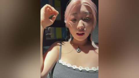 Media: Video of a young Asian woman with pale skin and pink hair, wearing a grey tank top with white lace trim. She has a surprised expression, with her left hand raised. The background is blurred, with a hint of a bookshelf.