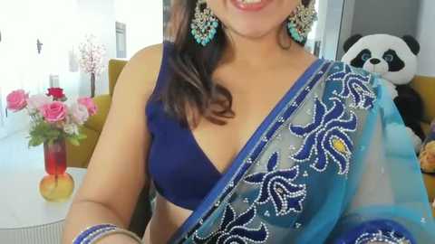 Video of a woman with medium brown skin, wearing a blue blouse and blue saree with ornate silver embroidery, standing in a modern living room with a panda stuffed toy, floral arrangement, and white walls.