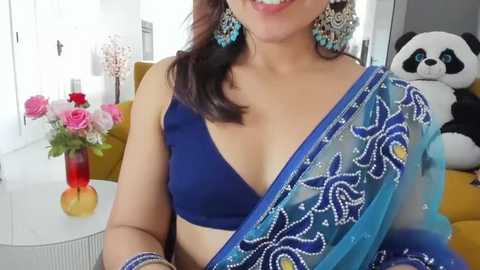 Media: Video of a smiling South Asian woman wearing a blue saree with intricate white floral embroidery, gold jewelry, and a dark blue bra. Background features a yellow couch, pink and red flowers in a vase, and a stuffed panda.