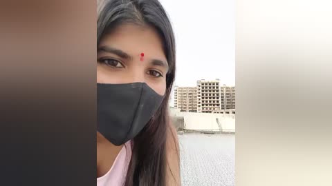 A close-up video of a South Asian woman with medium-brown skin, long black hair, and a red bindi, wearing a black face mask, taken outdoors near a river with multi-story buildings in the background.