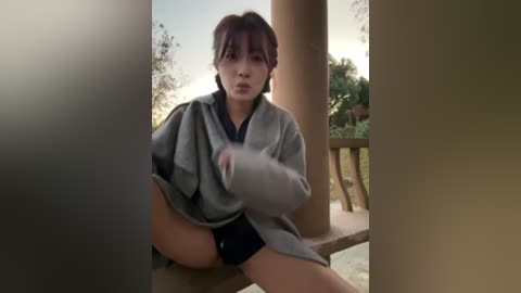 A video of an Asian woman with short brown hair, wearing a grey robe, sitting on a porch, with blurred background.