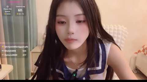 Video of an East Asian woman with long black hair, wearing a navy and white sailor-style top, sitting indoors. She has light skin and a neutral expression.