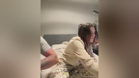 A video captures a young woman with shoulder-length brown hair, wearing a beige hoodie and patterned pajama pants, lying on a bed with a grey headboard. A tattooed man is seen behind her, possibly engaging in a sexual act. The room has a dimly lit, unmade bed and a wall-mounted light fixture.