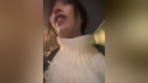 A blurred, close-up video of a woman with a turtleneck sweater, partially smiling, with brown hair. The background is out of focus.