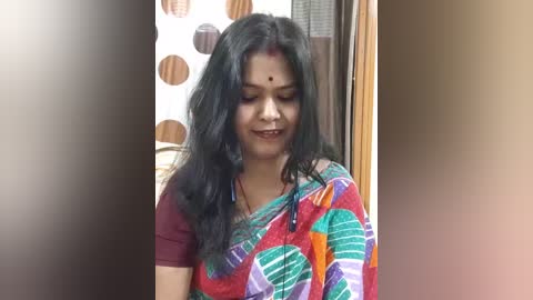 Video of a woman with long black hair, wearing a colorful sari with geometric patterns, applying mehndi on her forehead, in a domestic setting.