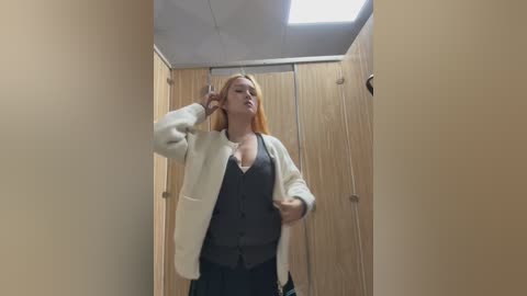 Video of a blonde woman in a beige cardigan and dark top, standing in a wooden-walled restroom, holding a phone to her ear, with a fluorescent ceiling light above.