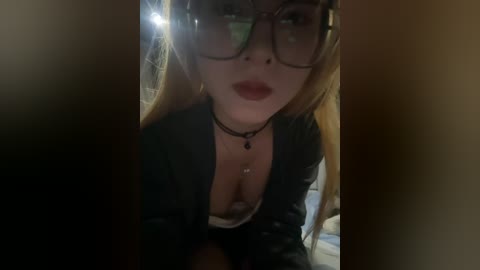 A dimly lit video shows a young woman with fair skin, long blonde hair, and large glasses, wearing a low-cut black top, revealing cleavage. She is indoors, likely in a bedroom, with soft lighting creating shadows.
