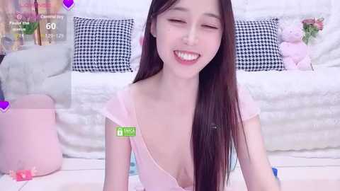Video of a young Asian woman with long dark hair, fair skin, and a slim build, smiling in a pink top. Background includes a white bed with black-and-white patterned pillows and stuffed toys.
