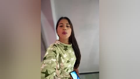 Video of a young Asian woman with long black hair, wearing a floral-patterned top, taking a selfie in a brightly lit room with soft, blurred background.