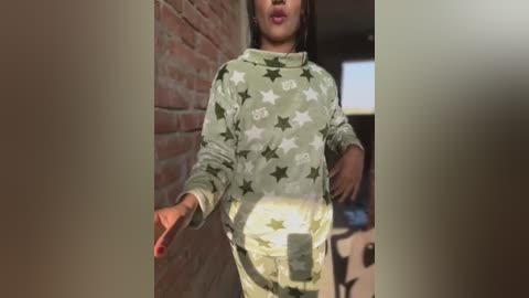 Video of a young person with medium-dark skin, wearing a camo sweatshirt with white stars, standing in a doorway with a brick wall and blurred interior behind them.