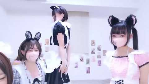Video of four Japanese women in black and white maid costumes, one with cat ears, standing in a minimalist room with white walls and a poster collage.