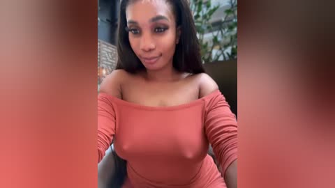 Video of a smiling Black woman with medium-dark skin tone, wearing a tight, off-the-shoulder, coral dress, with long black hair. Background features a blurred, modern indoor setting with plants.