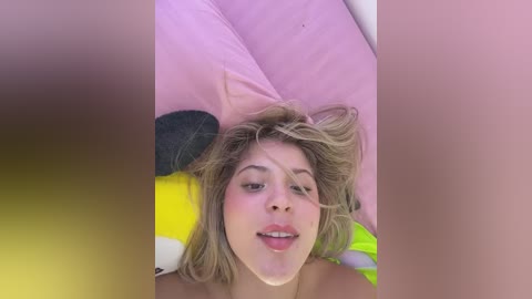 A video of a young woman with blonde hair lying on a pink pillow, wearing a yellow bikini top. The image is blurred, obscuring her face.