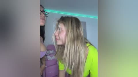 Video of a blonde woman with glasses in a bright green top, embracing a brunette woman in a purple top, both leaning against a white wall with green LED strip lighting.