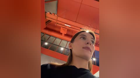 Video of a young woman with light skin and straight, brown hair, wearing a black top, standing in a modern, red-painted indoor space with industrial elements like exposed beams and ductwork.