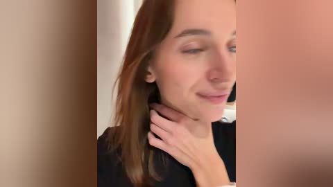 Media: A close-up video of a fair-skinned woman with long, straight brown hair, wearing a black top, gently touching her neck. She has a serene expression. The background is blurred.