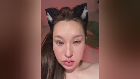 Video of a young woman with fair skin, wearing cat ears and minimal makeup, against a blurred pink and green background.