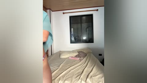 A video of a messy bedroom with a person wearing a blue shirt and white shorts, standing near a bed with beige sheets and a pink pillow.