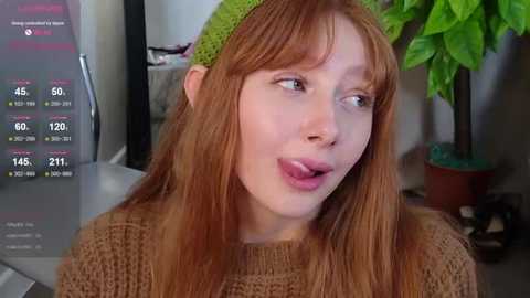 Video of a young Caucasian woman with long, straight red hair, wearing a green beanie and a brown knitted sweater. She appears to be in a modern, indoor setting with a plant in the background.