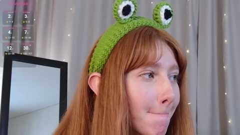Video of a smiling woman with long red hair, wearing a green knitted frog hat with large eyes, against a soft gray curtain backdrop with fairy lights.