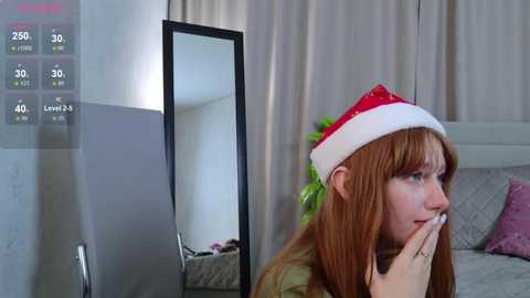 A video of a young woman with long, reddish-brown hair wearing a Santa hat, standing in a modern bedroom with a gray bed and beige curtains. She holds a phone to her mouth.
