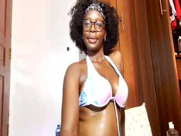 Video of a Black woman with a full Afro, wearing glasses and a tie-dye bikini, standing in a wooden closet.