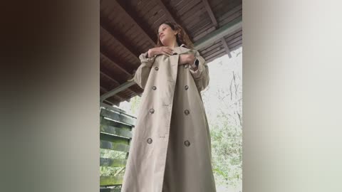 Video of a woman in a beige trench coat, standing under a wooden porch with greenery in the background. She appears relaxed, with a soft expression.