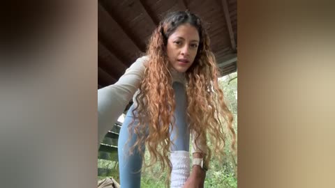 Video of a woman with long, wavy, caramel-colored hair, wearing a light blue top and white pants, bending forward, taken outdoors under a wooden roof.