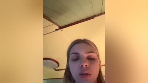 Video of a young woman with long, straight blonde hair, eyes closed, and mouth slightly open, standing in a room with yellow walls and a wooden ceiling.