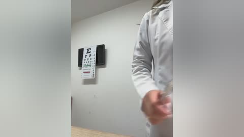 A video of a person in a white lab coat with a blurred hand, standing in front of a wall-mounted eye chart and wooden floor in a clinical setting.