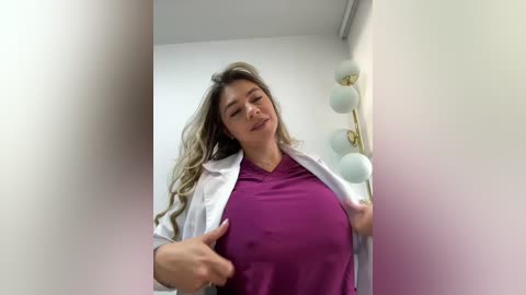 Video of a smiling, light-skinned woman with long, wavy blonde hair, wearing a purple top and open white lab coat, standing in a modern room with white walls and a golden lamp.