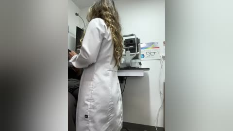 Video of a white female optometrist in a white lab coat, with long wavy brown hair, examining a patient in an optometry clinic.