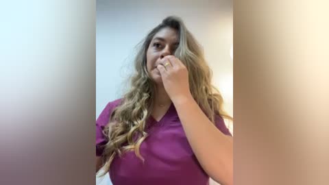 Video of a light-skinned woman with long, wavy blonde hair, wearing a purple shirt, covering her mouth with her hand, and standing in a softly lit, minimalist room with neutral walls.