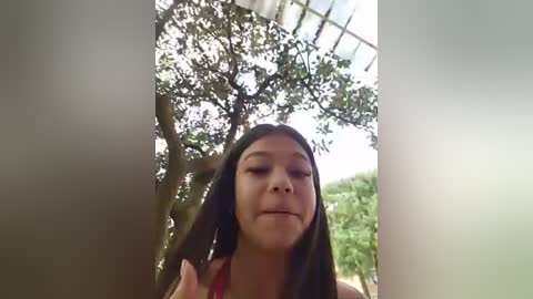 A video of a young Asian woman with long black hair and light skin, wearing a red top, standing in a sunlit, arched greenhouse with lush green trees in the background.