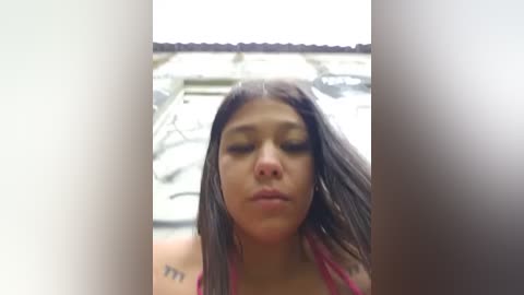 A video of a young woman with medium brown skin and long black hair, wearing a red tank top, looking downward with closed eyes. The background is blurry, possibly an indoor setting.