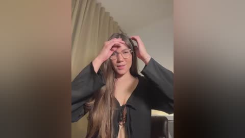 A video of a woman with long, straight brown hair, fair skin, and glasses, adjusting her hair. She wears a black blouse and is indoors with beige curtains in the background.