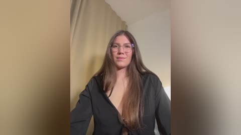A video of a smiling, fair-skinned woman with long, straight brown hair, wearing glasses and an open black shirt, standing against beige and cream-colored curtains.