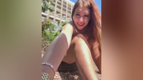 Video of a smiling, light-skinned woman with long brown hair, wearing a tank top, shorts, and silver bracelets, sitting on the ground outside near a multi-story building.