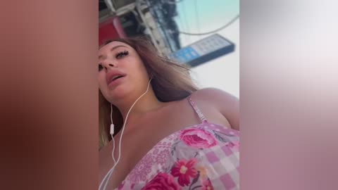 Video of a young woman with light skin and long, wavy brown hair, wearing a floral-patterned top, listening to music on white earbuds. Background shows a cluttered room with electrical cords and a blue sign.