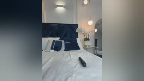 Video of a modern bedroom with a white bed, dark blue pillows, and a tufted headboard. White walls, a round mirror, and a vase of white orchids are visible.