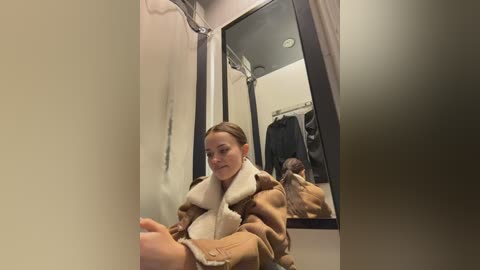 A video of a woman with light skin and brown hair, wearing a beige fur coat, sitting in a dressing room with a large mirror reflecting her and a black coat hanging behind her.