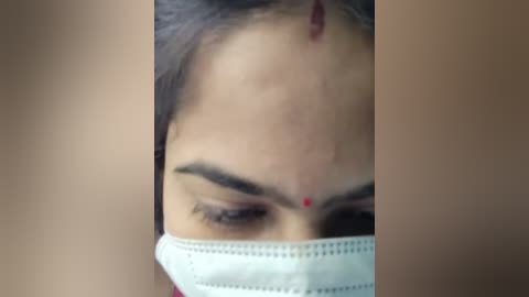 A close-up video of a South Asian woman with medium brown skin, wearing a white surgical mask, showing a red dot on her forehead, and dark hair. The background is blurred, focusing on her face.